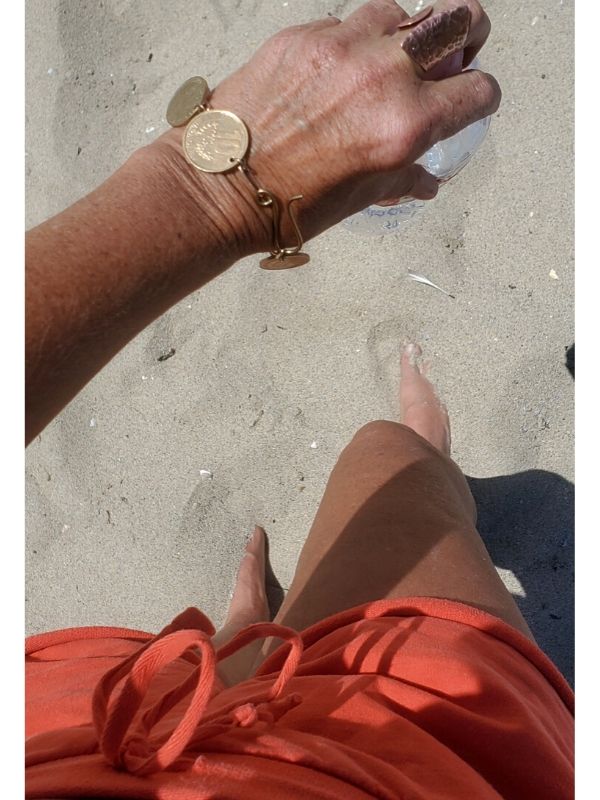 at the beach in orange shorts and a gold coin brACELET